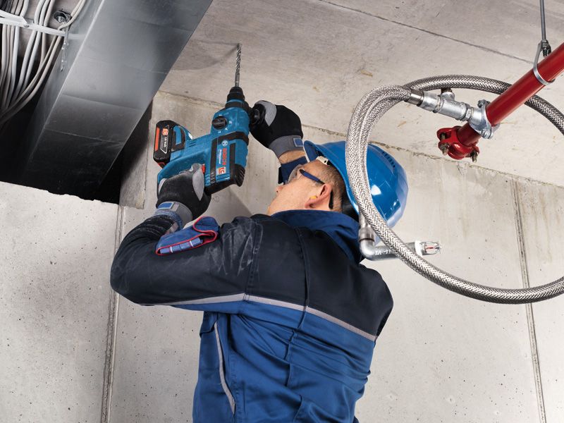 A person wearing safety equipment drills into a concrete ceiling with a cordless rotary hammer.