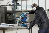 A person wearing safety equipment cuts a metal pipe with a metal cut-off saw.