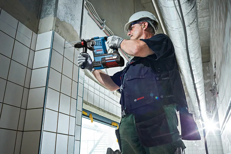 A person wearing safety equipment drills into tiled wall using a cordless rotary hammer.