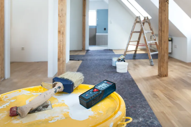 Paintbrush and laser measure on a yellow paint bucket in a room under renovation.