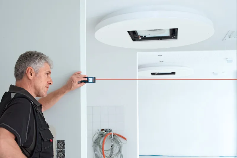A person uses a laser measure to check distance across a white room under construction.