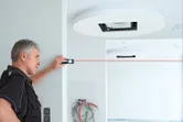 A person uses a laser measure to check distance across a white room under construction.
