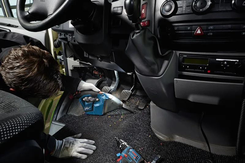 A person wearing safety equipment cleans a car floor with a cordless vacuum cleaner.