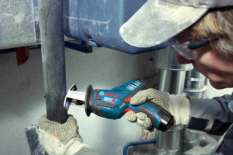 A person wearing safety equipment cuts a plastic pipe with a cordless reciprocating saw.