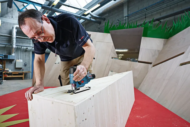A person uses a cordless jigsaw to cut wood in a workshop.