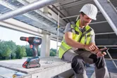 建築工地上，一名配戴安全設備的工人在無線電鑽旁使用智慧型手機。
