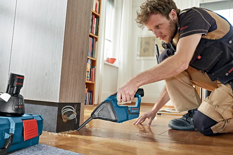 A person wearing safety equipment uses a cordless vacuum cleaner to clean wood shavings.