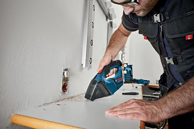 A person wearing safety equipment uses a cordless vacuum cleaner to clean a dusty surface.