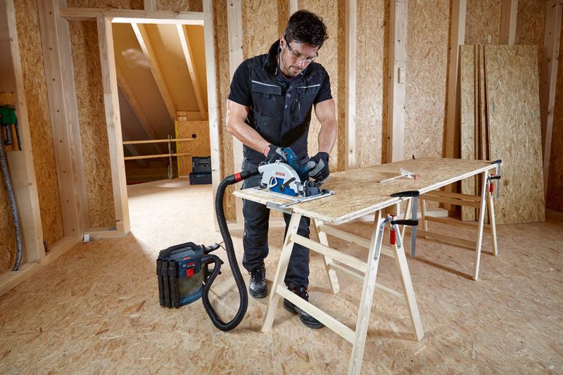 一名配戴安全設備的人員使用連接到除塵器的圓鋸切割膠合板。.