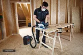 A person wearing safety equipment cuts plywood with a circular saw connected to a dust extractor.