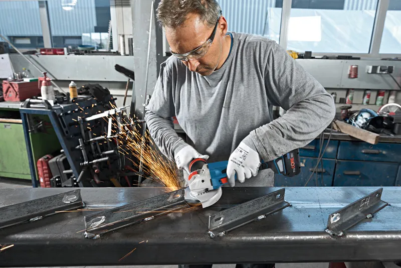 A person wearing safety equipment grinds metal with a cordless angle grinder, producing sparks.