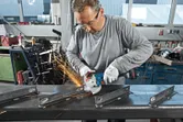 A person wearing safety equipment grinds metal with a cordless angle grinder, producing sparks.
