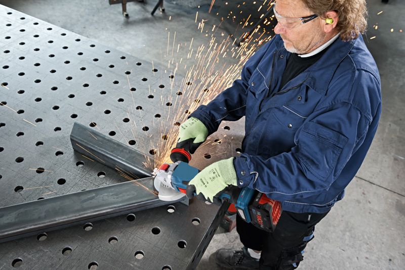 A person wearing safety equipment grinds metal beams with a cordless angle grinder.
