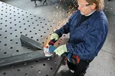 A person wearing safety equipment grinds metal beams with a cordless angle grinder.