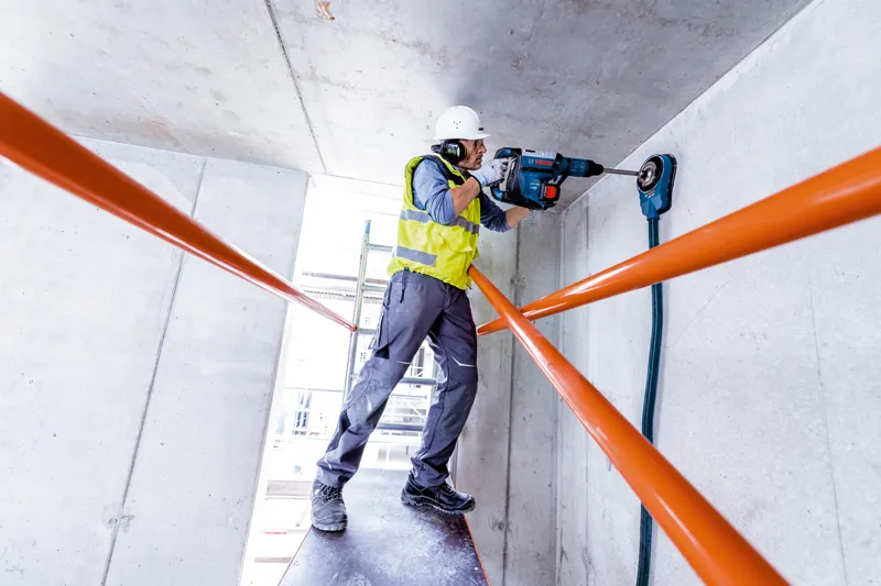 一名配戴安全設備的人員使用旋轉錘在混凝土天花板上鑽孔。.