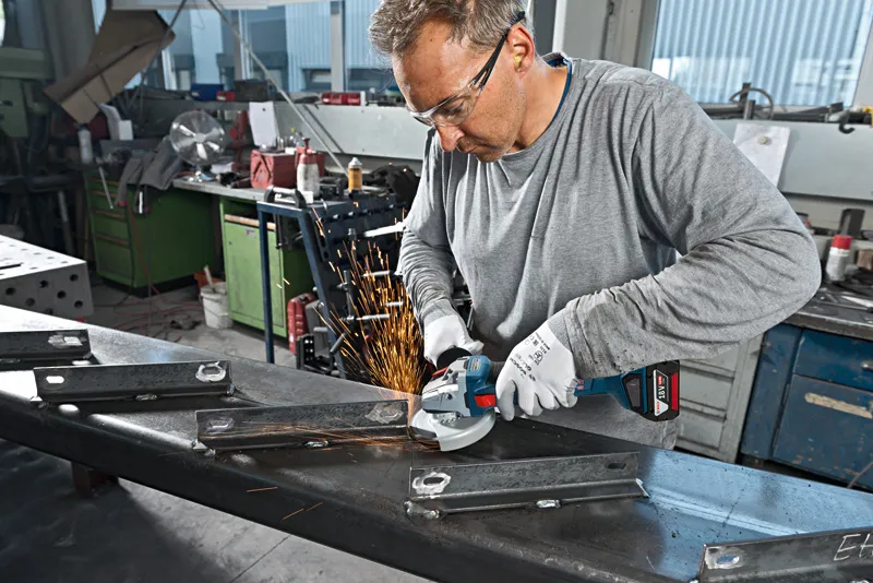A person wearing safety equipment grinds metal beams with an angle grinder, producing sparks.