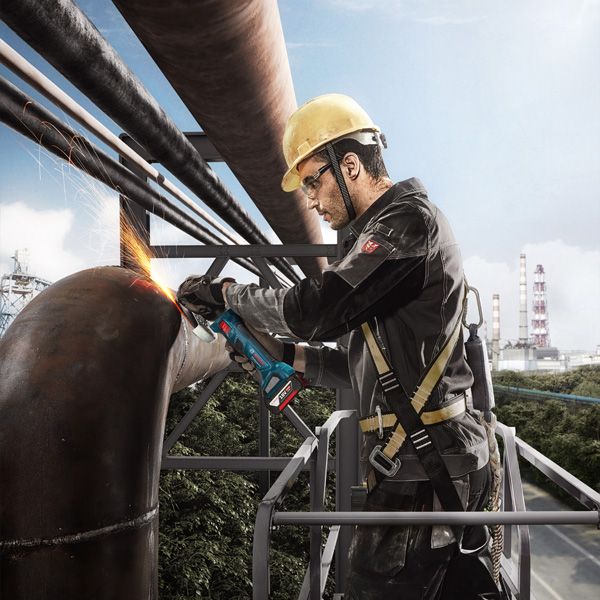 A person wearing safety equipment grinds a large metal pipe with an angle grinder.