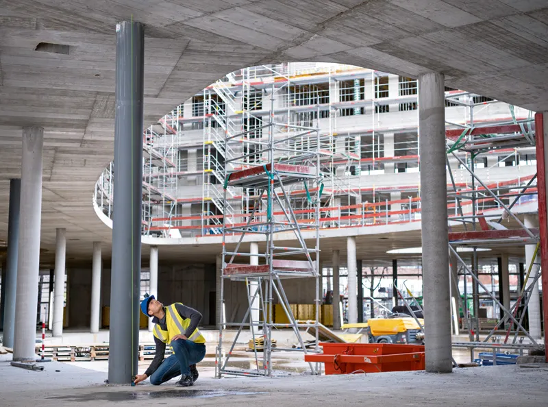 A person wearing safety equipment uses a laser measure to check alignment at a construction site.