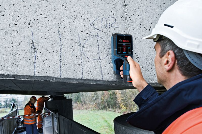 A person wearing safety equipment uses a detector on a concrete beam.