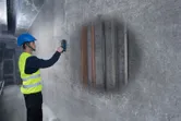 A person wearing safety equipment uses a detector to scan for pipes and rebar inside a wall.