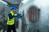 A person wearing safety equipment scans a concrete wall with a detector showing internal structures.