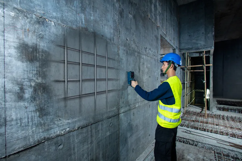 A person wearing safety equipment scans a concrete wall with a detector showing embedded rebar.