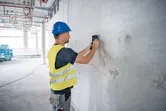 A person wearing safety equipment scans a concrete wall with a detector.