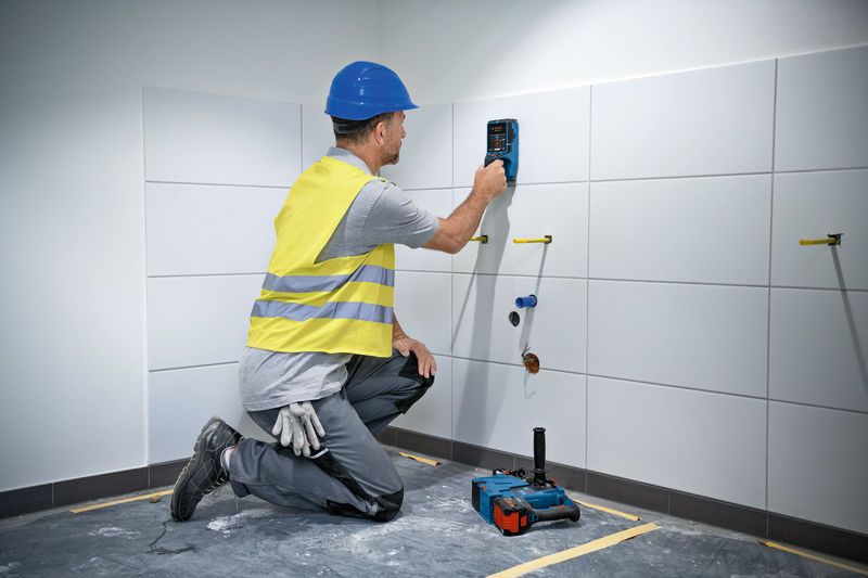 A person wearing safety equipment uses a wall scanner on a tiled wall.