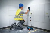 A person wearing safety equipment uses a wall scanner on a tiled wall.