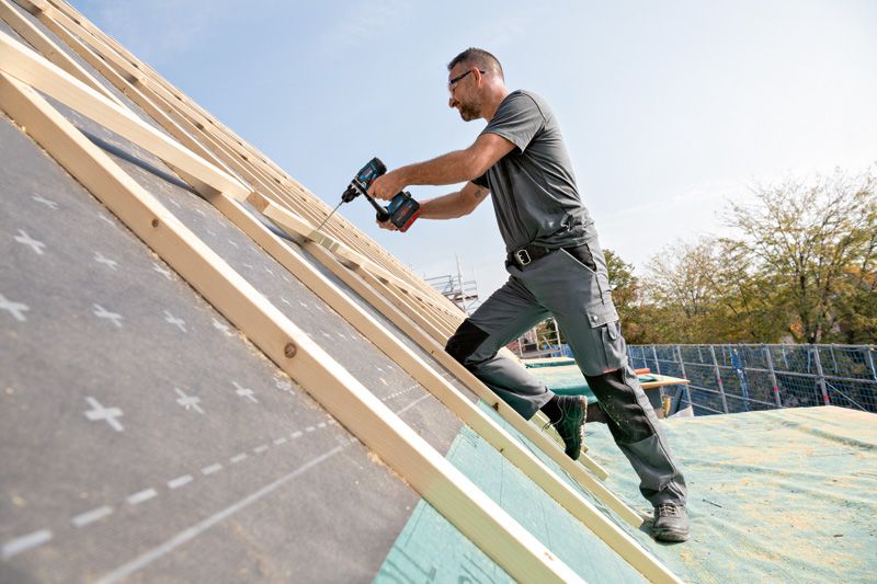 GSR 18V-150 C 一名佩戴安全設備的人員使用無線電鑽將木頭固定在傾斜的屋頂上。.