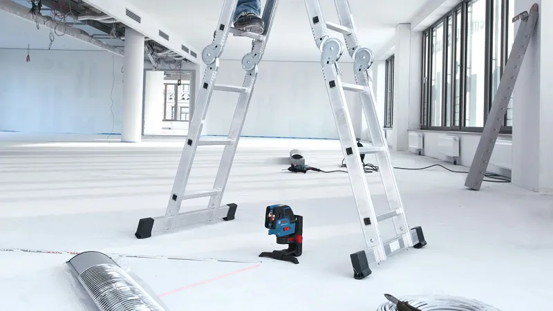 A laser leveling tool projects red lines on the floor in a bright, unfinished room.