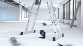 A laser leveling tool projects red lines on the floor in a bright, unfinished room.