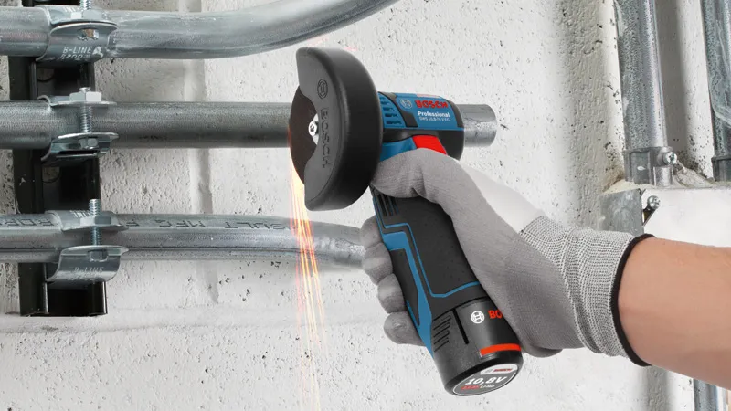 A person wearing safety equipment cuts metal conduit with a cordless angle grinder.