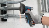 A person wearing safety equipment cuts metal conduit with a cordless angle grinder.