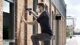 Person wearing safety equipment drills into a brick wall outside a building.