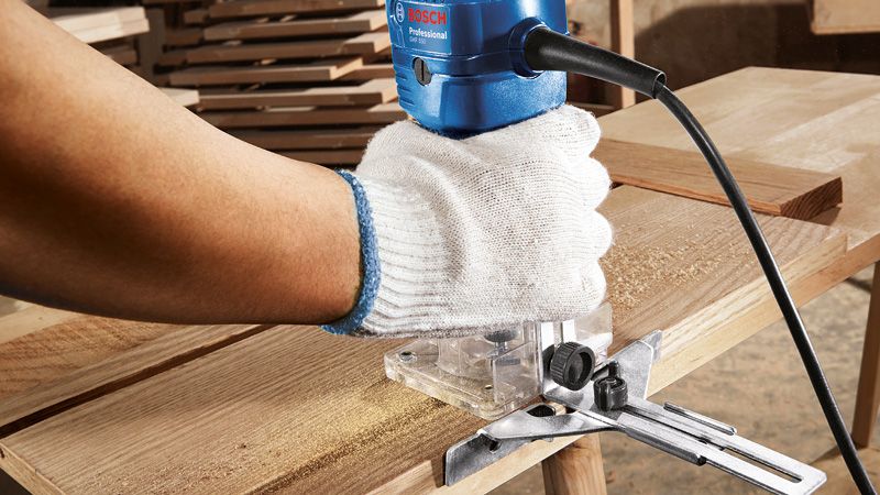 A person wearing safety equipment trims wood with a palm router on a workbench.
