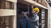 A person wearing safety equipment drills into a wooden beam at a construction site.