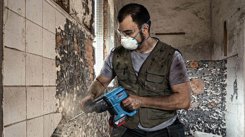 A person wearing safety equipment removes tiles from a wall using a power tool.