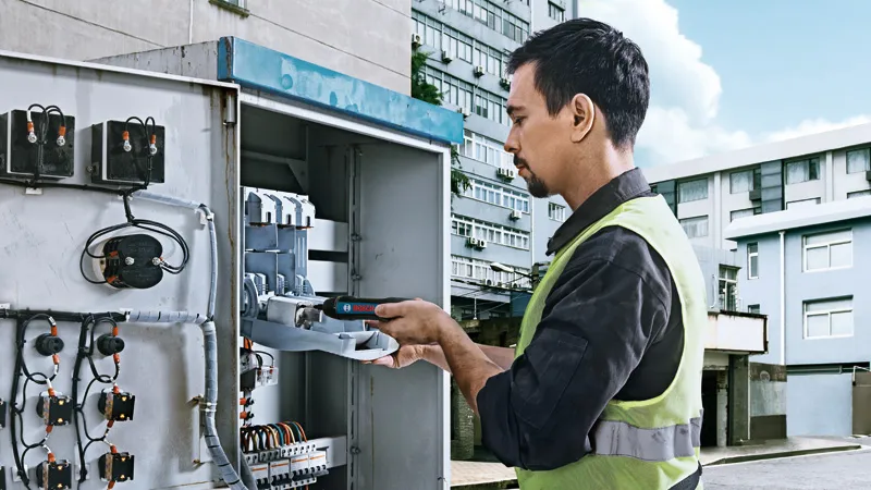 一名配戴安全設備的人員正在使用無線螺絲起子操作電氣面板。.