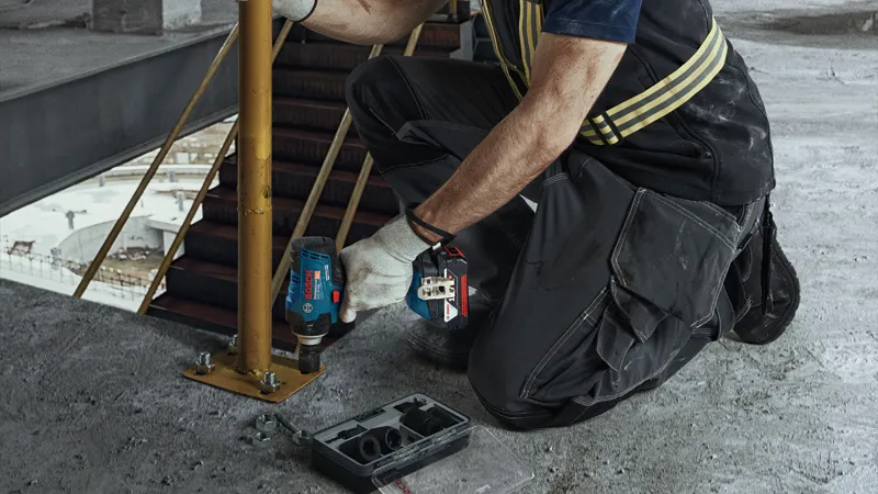 一名配戴安全設備的人員使用無線衝擊扳手來擰緊金屬柱上的螺栓。.