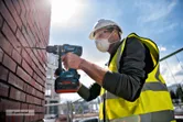 一名穿戴安全裝備的工人正在建築工地用電鑽鑽磚牆。