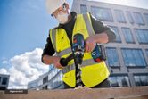 A person wearing safety equipment drills into wood with a cordless drill at a construction site.