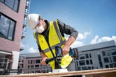 A person wearing safety equipment drills a hole into wood at a construction site.
