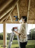 A person on a ladder drills into a wooden roof frame, wearing safety equipment.
