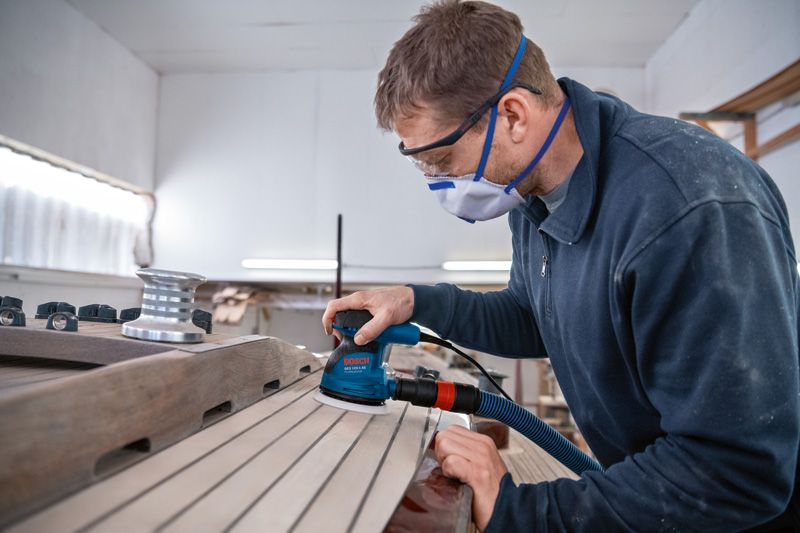 一名配戴安全設備的人員正在使用隨機軌道砂光機打磨木板。.