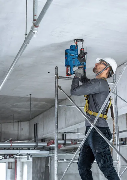 A person wearing safety equipment drills into a concrete ceiling while standing on scaffolding.