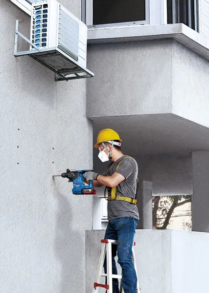 A person wearing safety equipment drills holes into a concrete wall beneath an air conditioner.