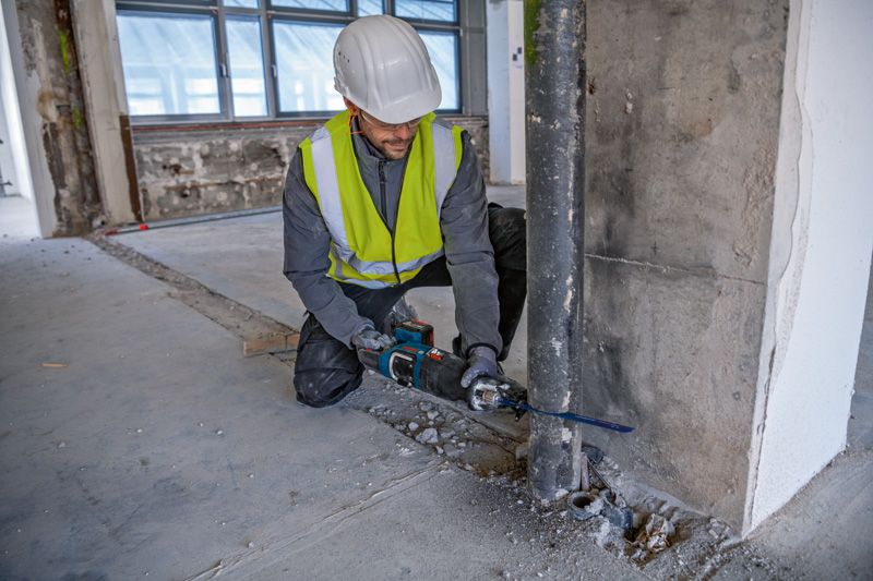 A person wearing safety equipment uses a reciprocating saw to cut a metal pipe.