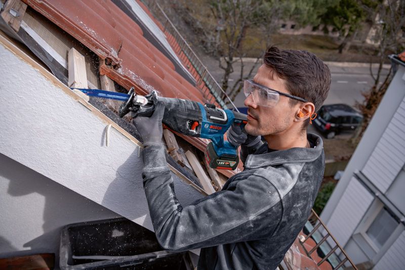 A person wearing safety equipment uses a reciprocating saw to cut wood on a rooftop.