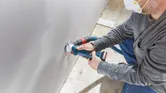 一名配戴安全設備的人員使用無線乾牆路由器在乾牆上開了一個洞。.